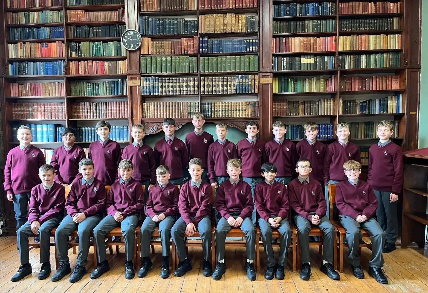 A class group of students in maroon jumpers photographed in the college library in front of the bookshelves